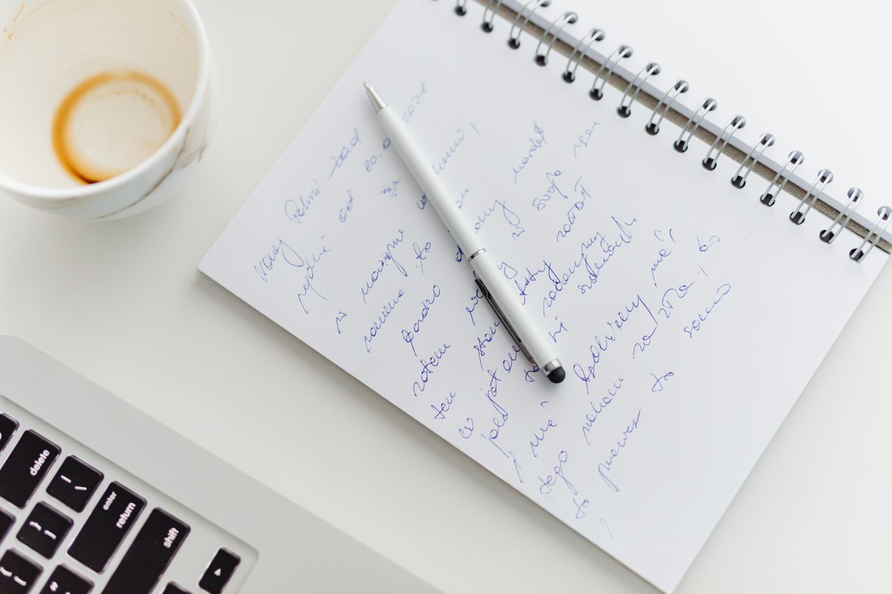 Close up of a notebook with handwriting, a pen, coffee cup, and laptop on a white desk.